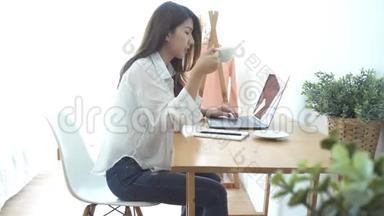 美丽的年轻微笑的女人，在笔记本电脑上工作，一边享受着温暖的咖啡，坐在家里的客厅里。